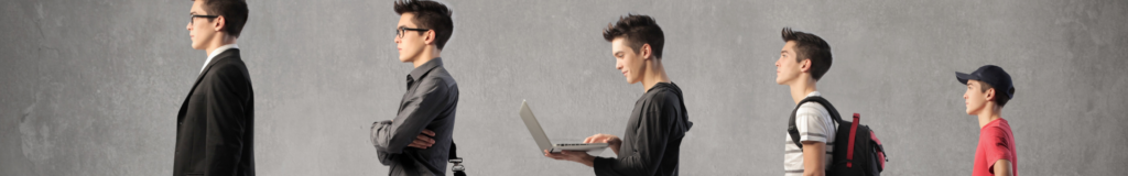A young man holding and using a laptop against a plain gray wall.