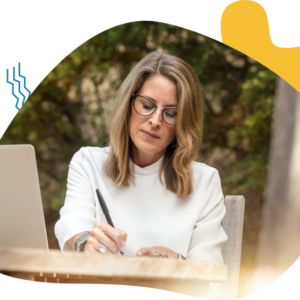 Woman writing notes outdoors with laptop nearby.